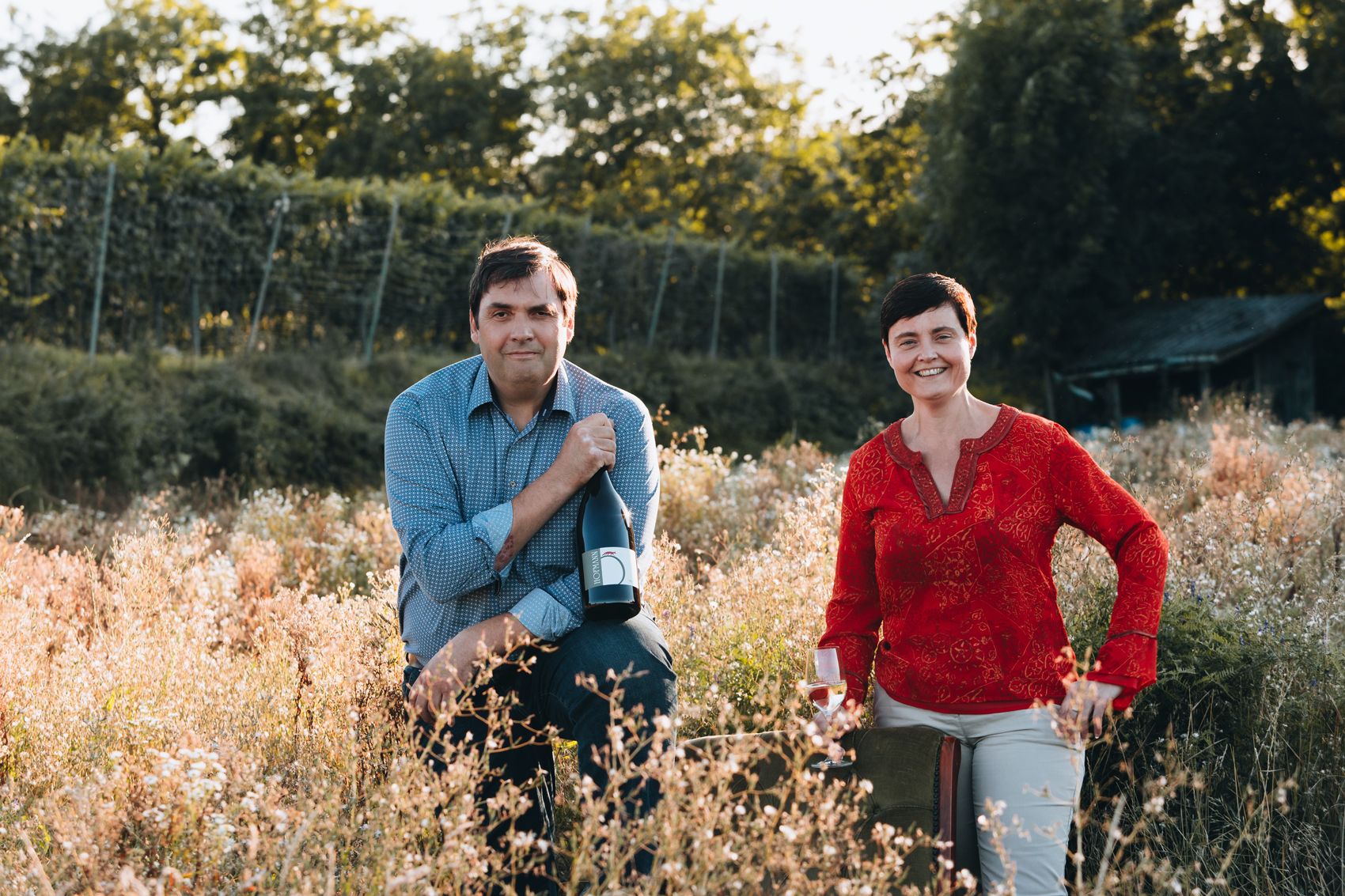 Two people in a vineyard, one holding a wine bottle, the other a wine glass.