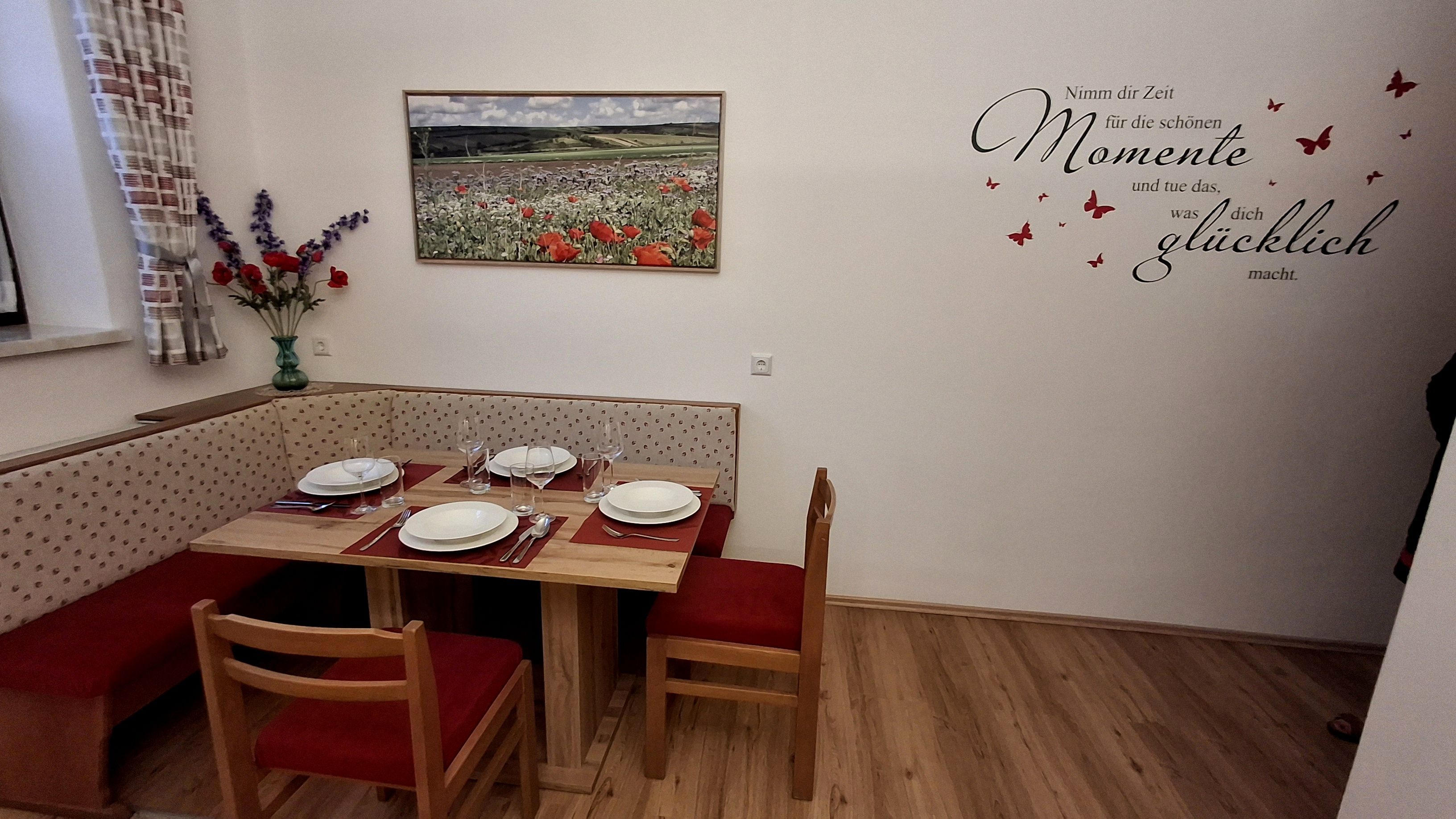 Dining area with corner bench, table and chairs, set with crockery. On the wall, a picture of poppies and a quote with butterflies.