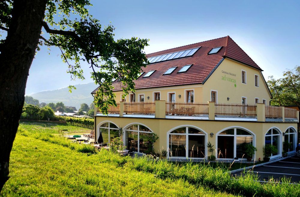Yellow building with red roof and terrace, surrounded by green landscape.