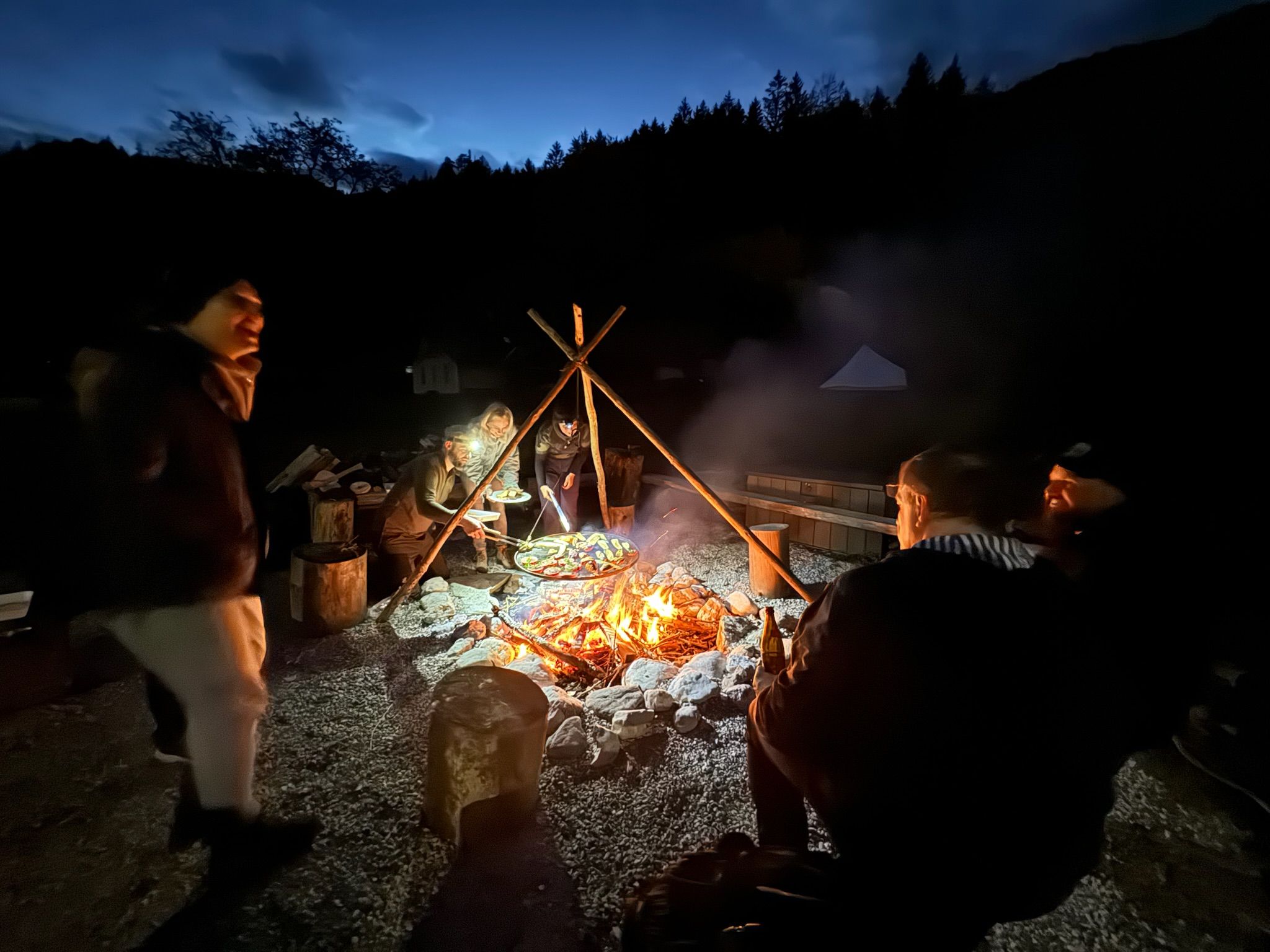 People sitting around an outdoor campfire at night, surrounded by trees.