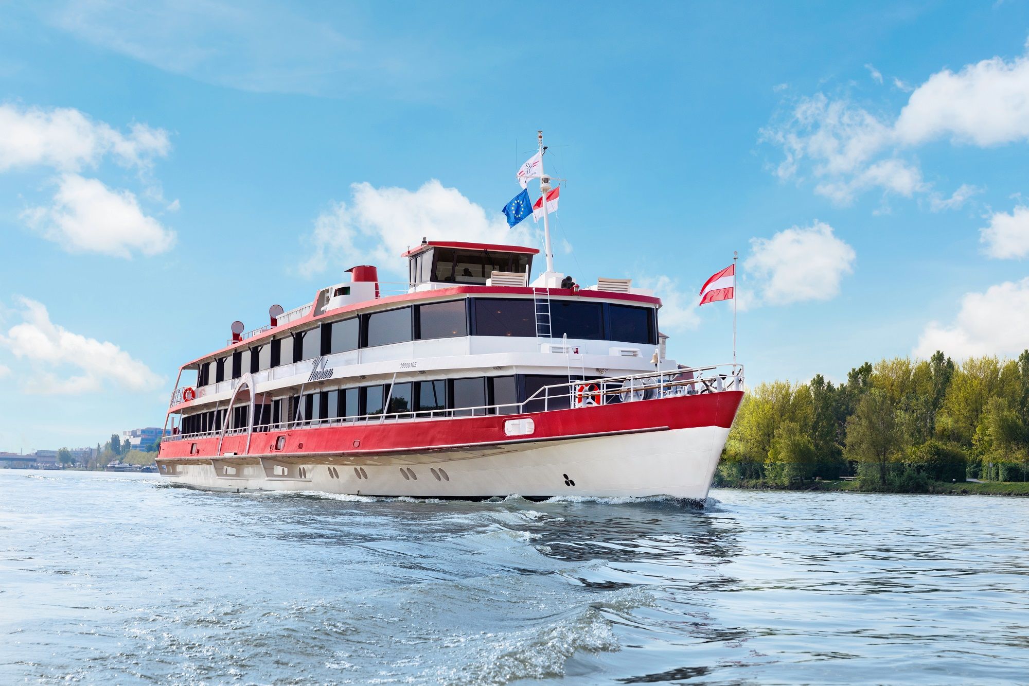 A passenger ship with red and white colors sails on a river under a blue sky.