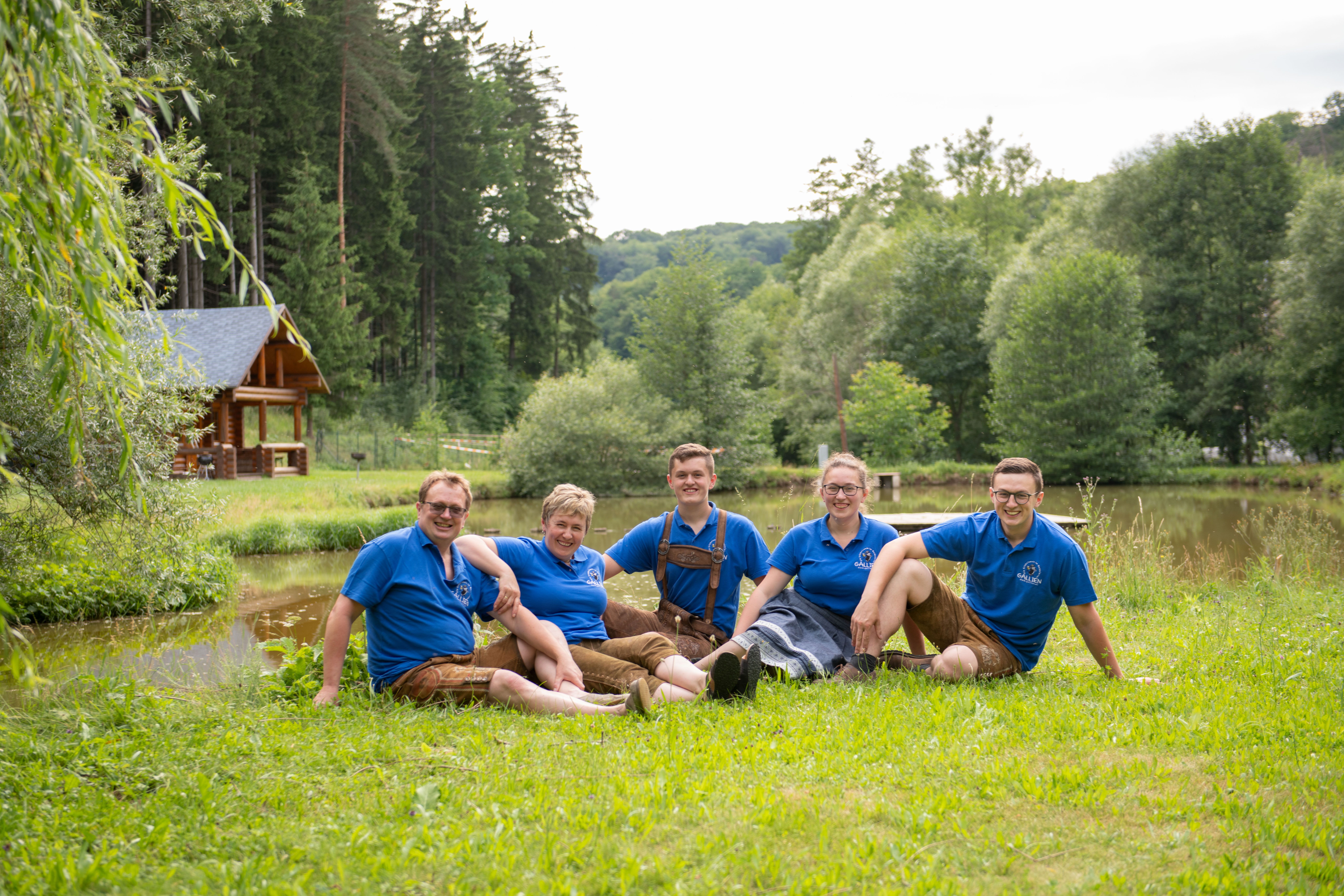 Five people wearing blue shirts are sitting on a lawn in front of a pond and a wooden house.