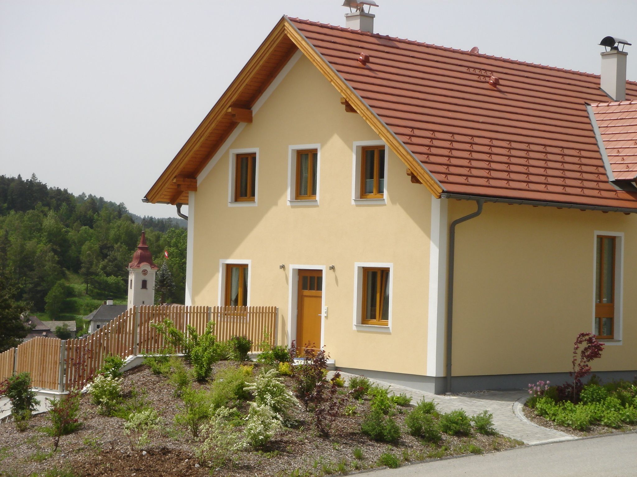 A modern yellow house with a red roof and garden, with a church and green landscape in the background.