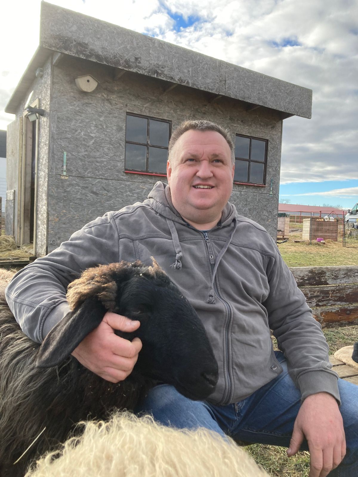A man is sitting outside next to a black sheep, which he is stroking. A small building can be seen in the background.
