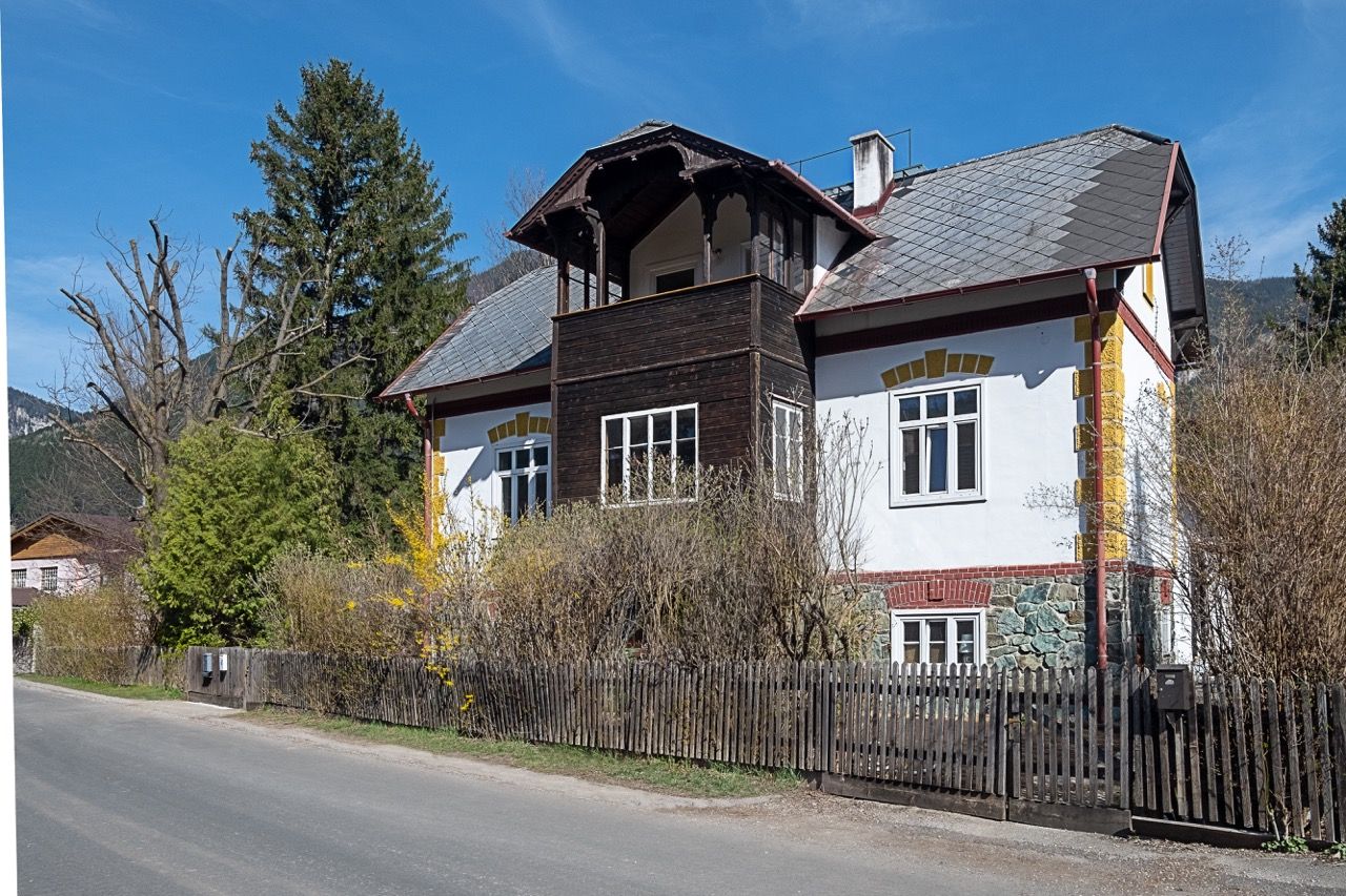 A traditional country house with a wooden balcony and white façade, surrounded by trees and shrubs, under a clear blue sky.