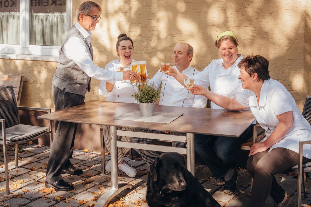 A team of five people clink glasses at a table outside, with a black dog in the foreground.