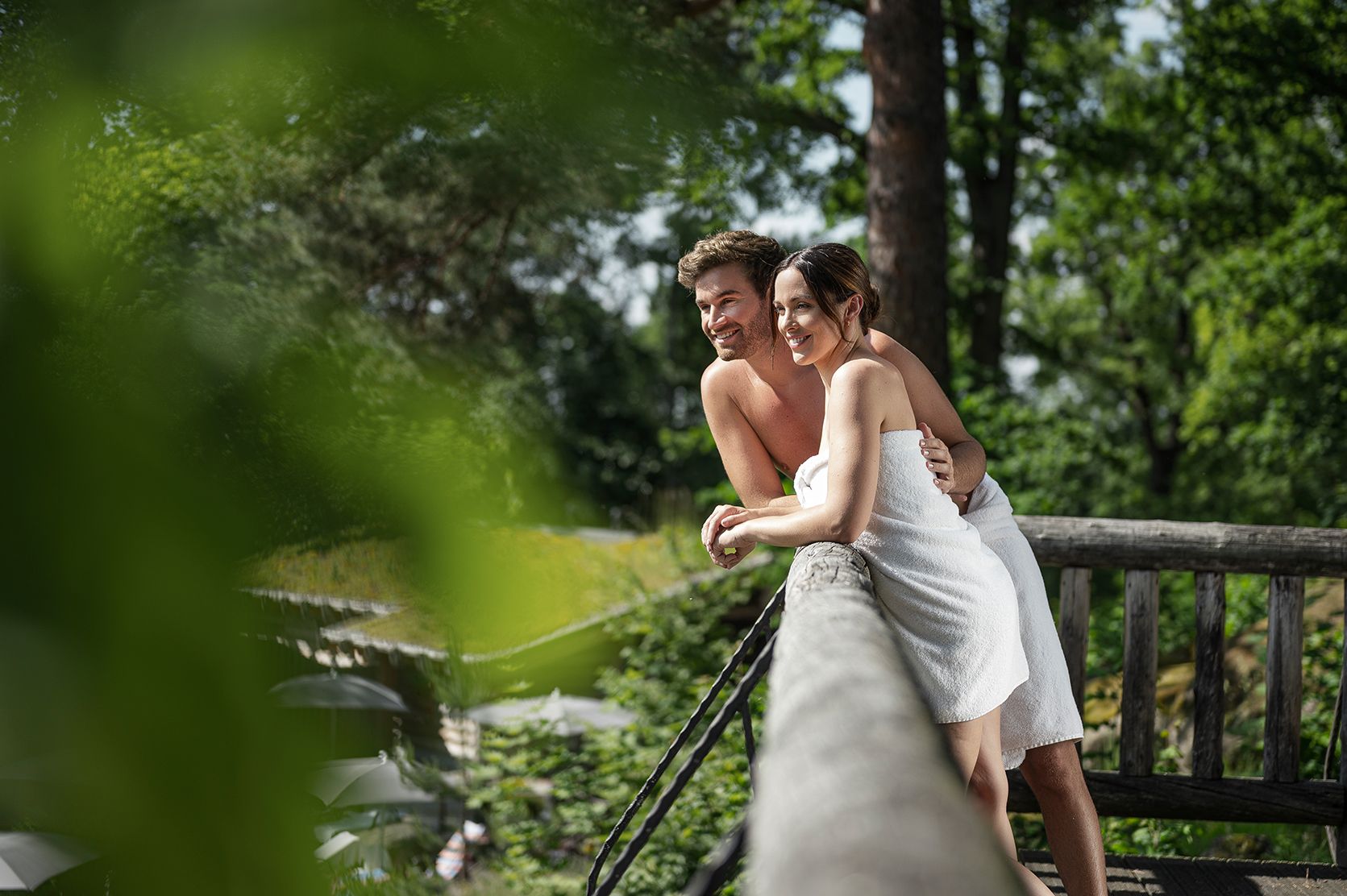 A couple in towels lean smiling against a railing outside, surrounded by green trees.