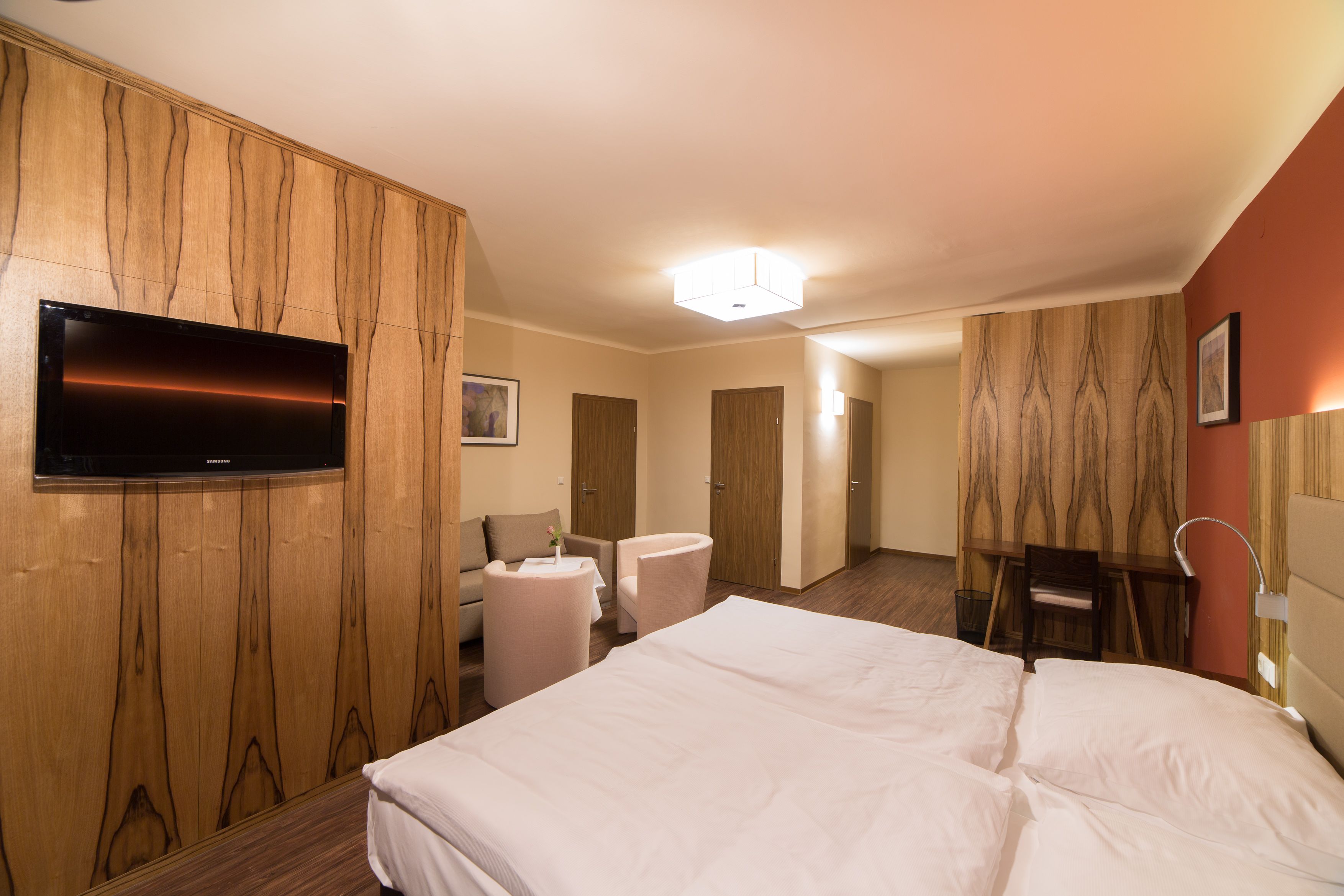 Modern hotel room with double bed, TV, seating area and wooden furniture.