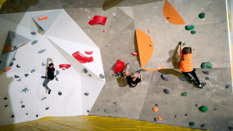 Three children climbing on an indoor bouldering wall with colorful holds.