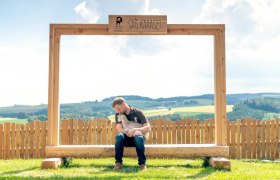 A man sits on a wooden bench under a large frame with the inscription 'SAG KÄÄÄSE' and holds a kid. A rural landscape can be seen in the background.