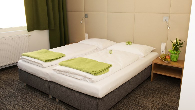 Comfortable hotel room with double bed, green towels and bedside table with flowers and fruit.