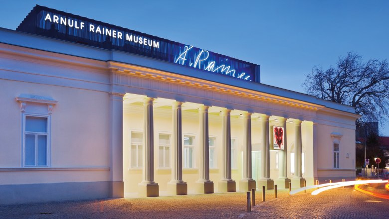 Exterior view of the Arnulf Rainer Museum at dusk with illuminated columns and neon sign.
