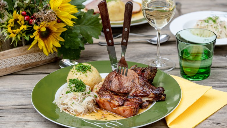 A plate of roast pork, dumplings and sauerkraut on a wooden table, next to it a glass of white wine and a flower arrangement.