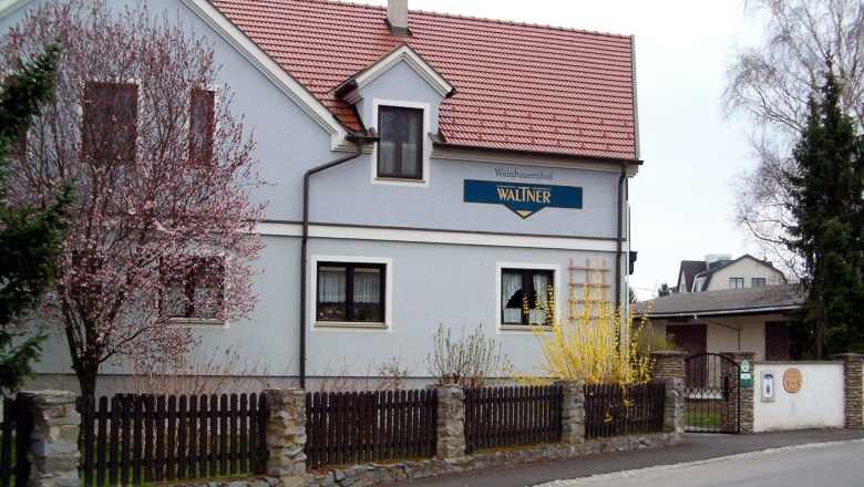 House, © Waltner A two-storey house with a red roof and blue façade, surrounded by blossoming trees and a wooden fence.