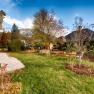 An autumnal garden with trees, benches and a paved path. Mountains and a blue sky can be seen in the background.
