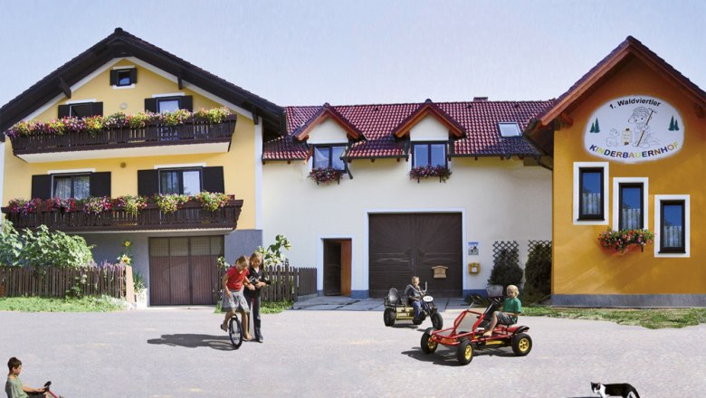 Children play on Kettcars in front of a yellow farm with flower balconies.
