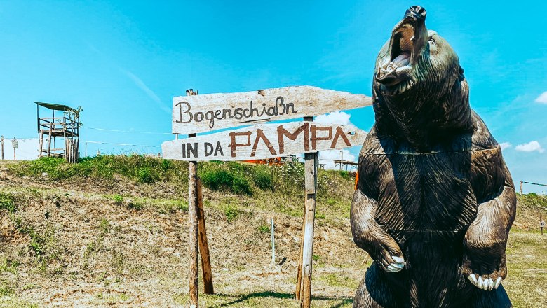 A large bear model next to a sign with the inscription 'Bogenschia&szlig;n in da Pampa' on a meadow.