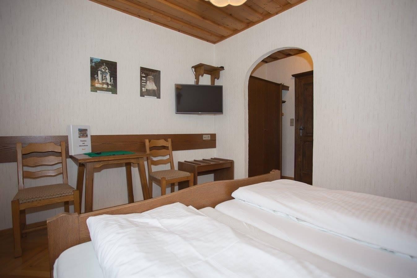 A cozy double room with wooden furniture, a double bed, a small table with two chairs and a TV on the wall.