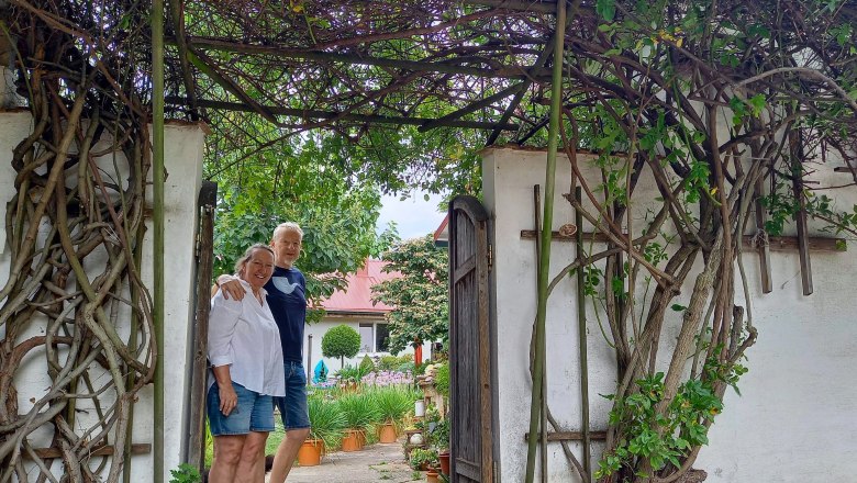 Two people stand smiling under an archway overgrown with plants in a garden.