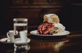 A plate with a monastery cream slice, garnished with red berries, stands on a wooden table. A glass of water and two espresso cups can be seen in the background.