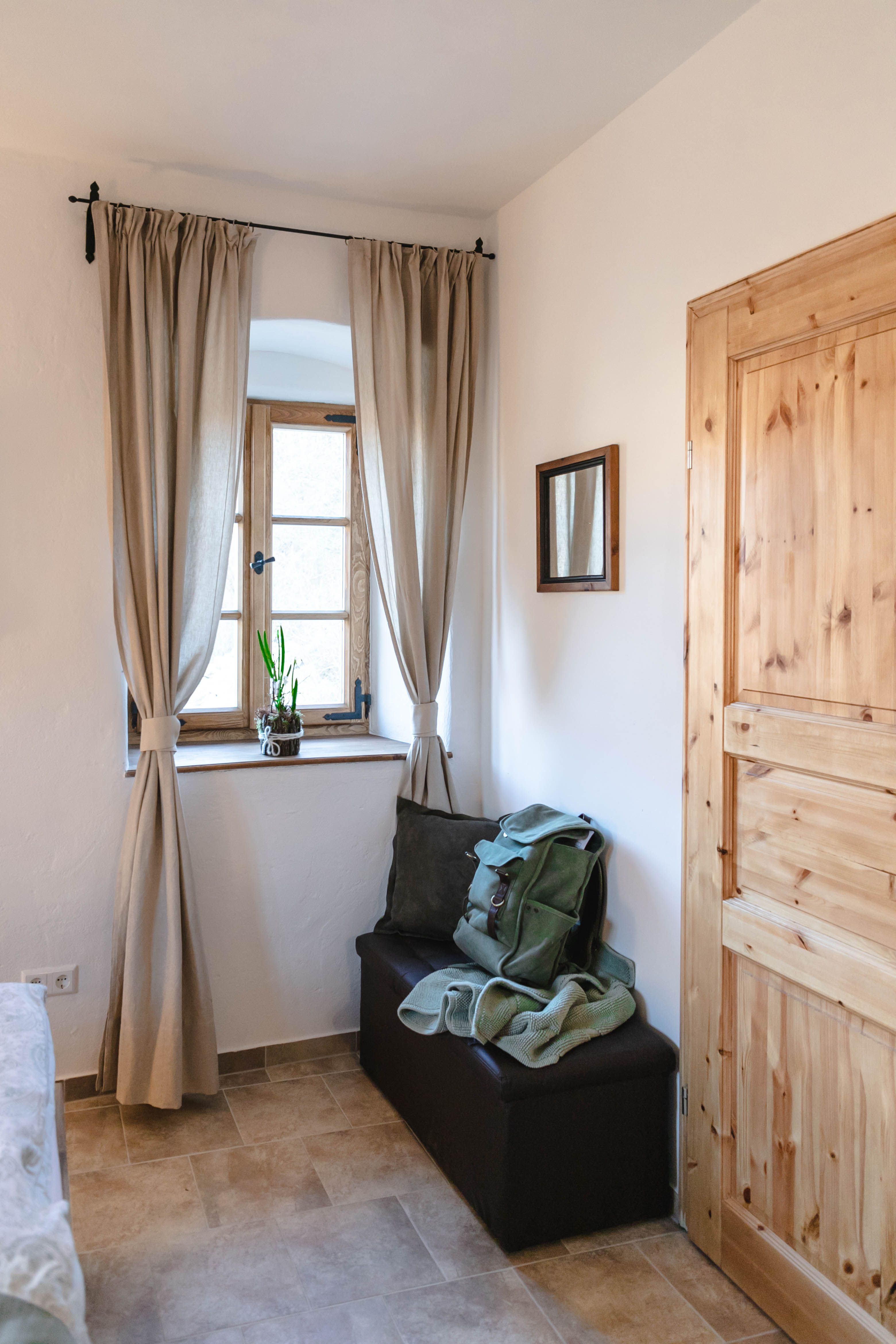 A small anteroom with a window, curtains, a wooden door and a bench with a rucksack and blanket.