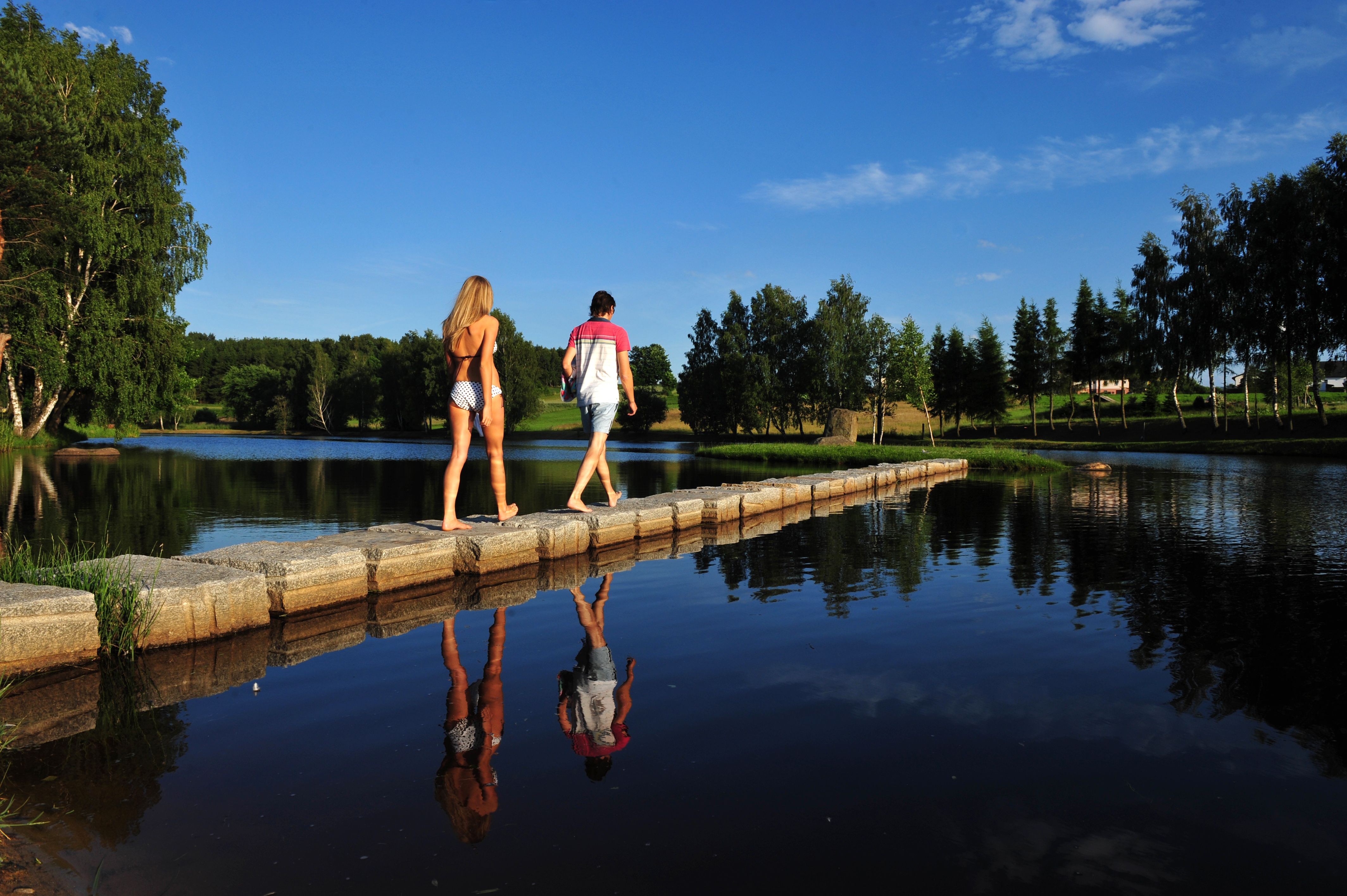 Two people walk on a footbridge over a lake in the Schrems moor spa, surrounded by trees and blue sky.