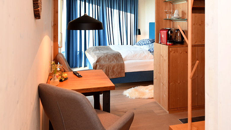 Cozy hotel room with wooden furniture, a bed, table and chairs. There is a coffee machine on a shelf. Blue curtains at the window.