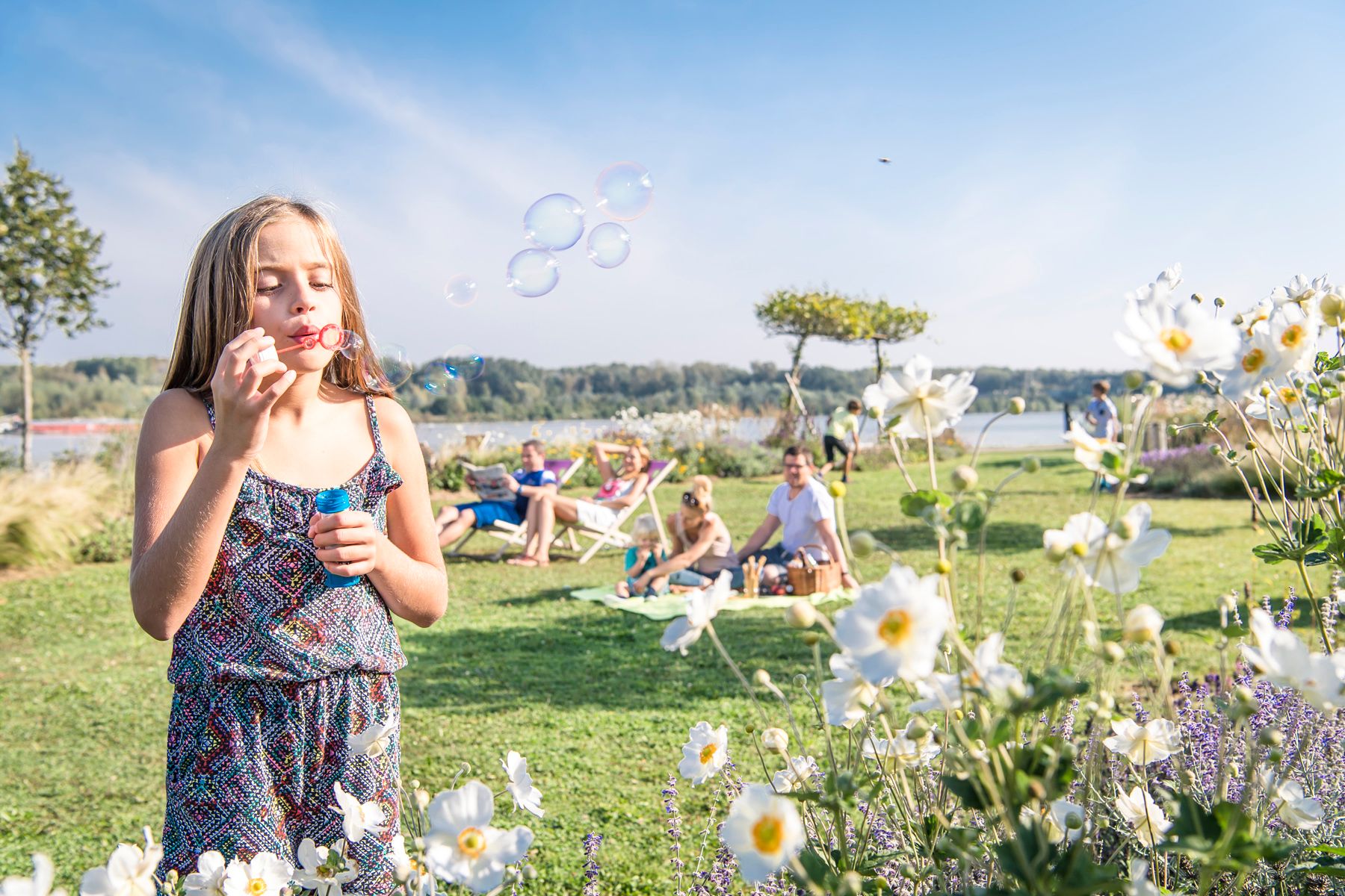 Girl making soap bubbles in a park with flowers and people in the background.