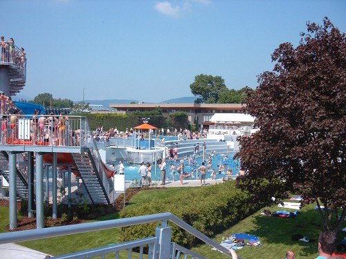 A lively outdoor pool with lots of people, water slides and sunbathing lawn.