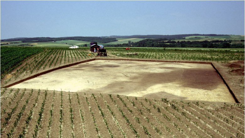 Archaeological excavation site in a field with agricultural machinery in the background.