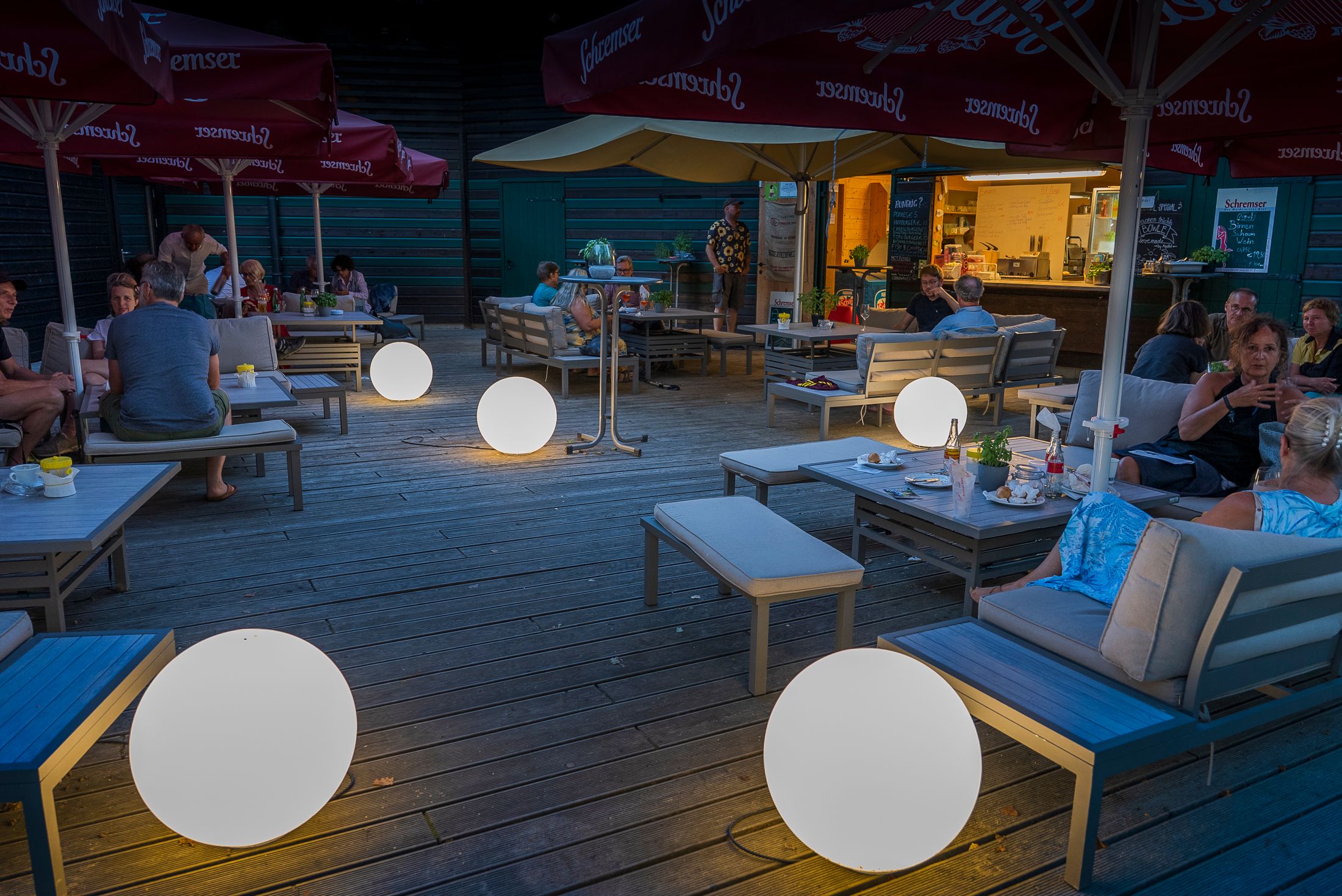 People are sitting in a cozy lounge by the lake, surrounded by glowing balls and umbrellas.