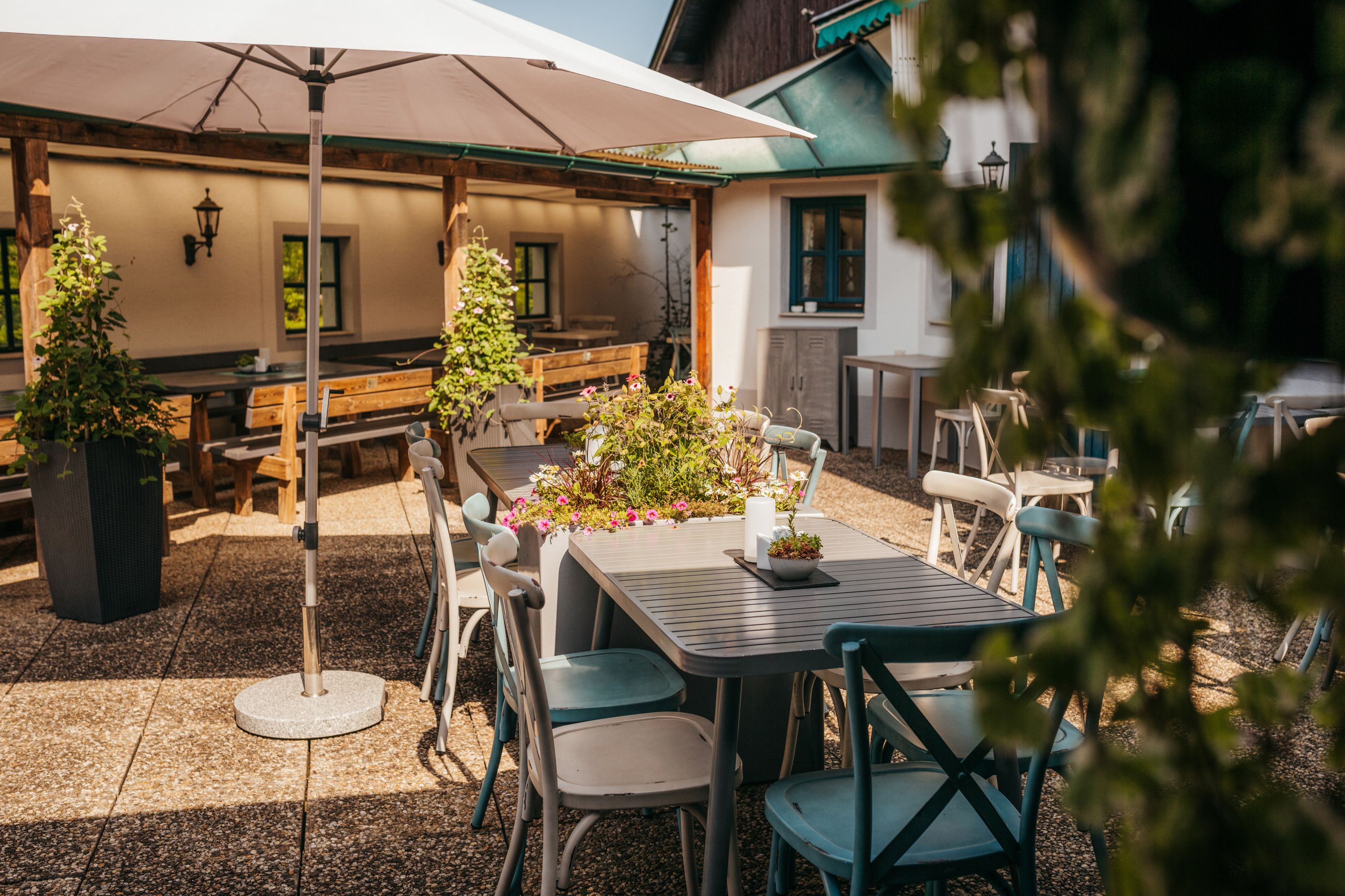 A sunny terrace with tables, chairs and plants under a parasol.