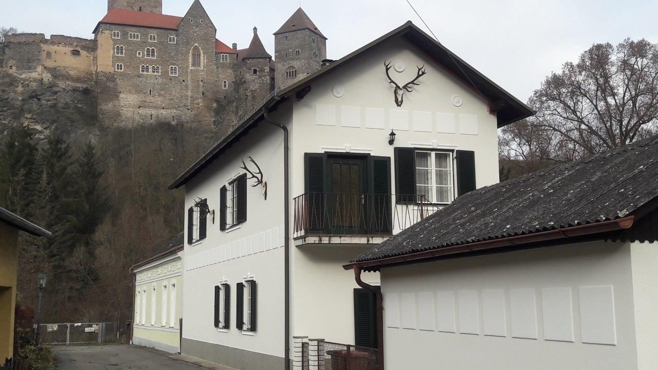 A white house with green shutters and deer antlers on the wall, a castle on a hill in the background.