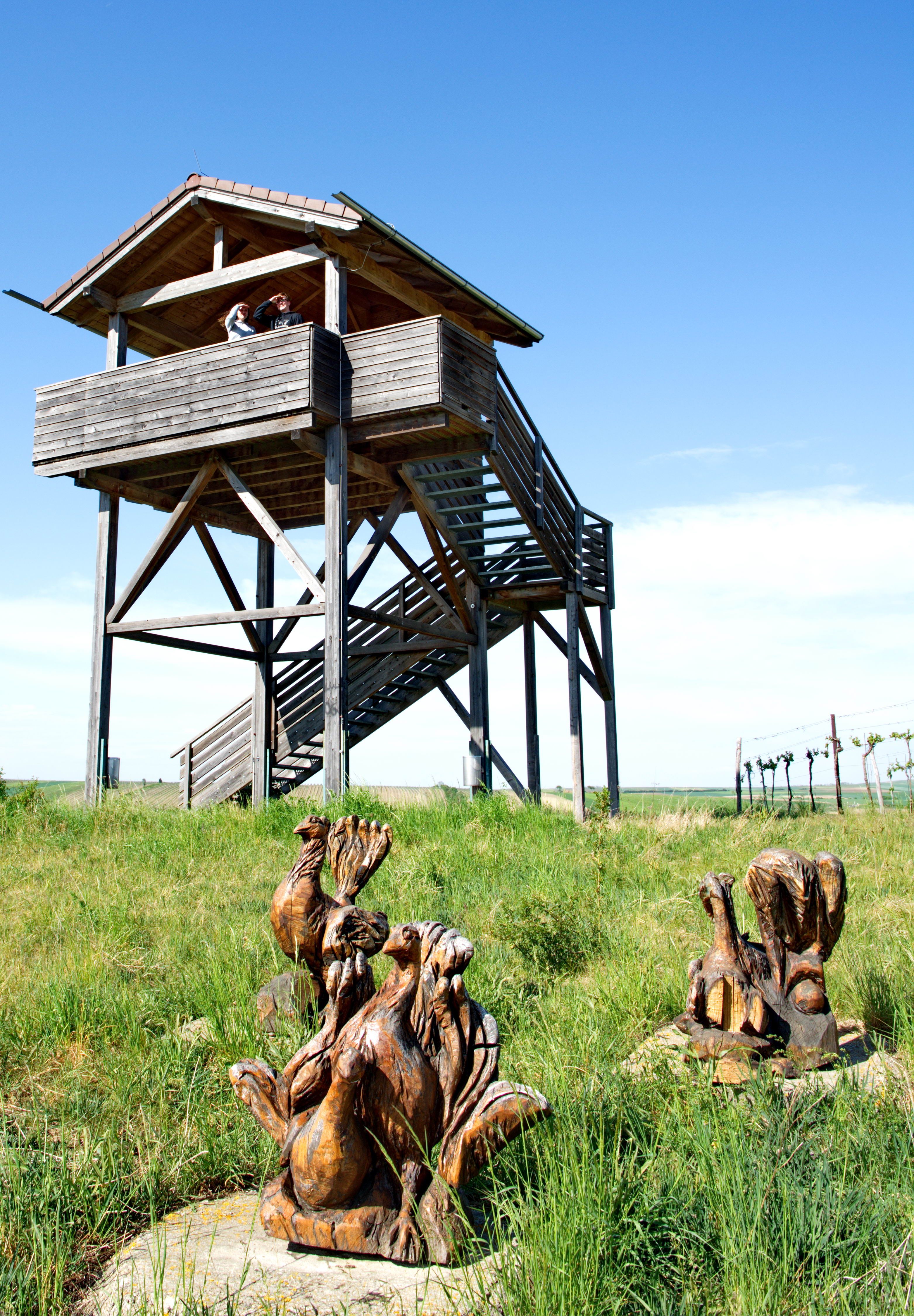 Wooden tower with two people and wooden sculptures of birds in the foreground on a meadow.