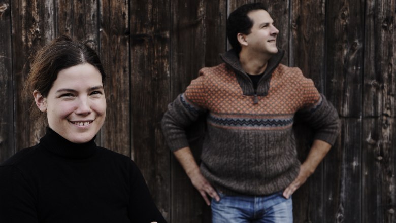 Two people in front of a wooden wall, a woman in the foreground smiling, a man in the background looking to the side.