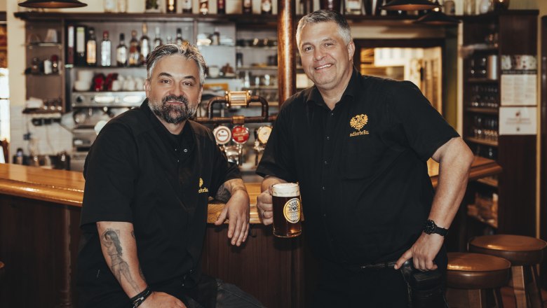 Two men dressed in black are standing in a bar with a beer mug in their hands.