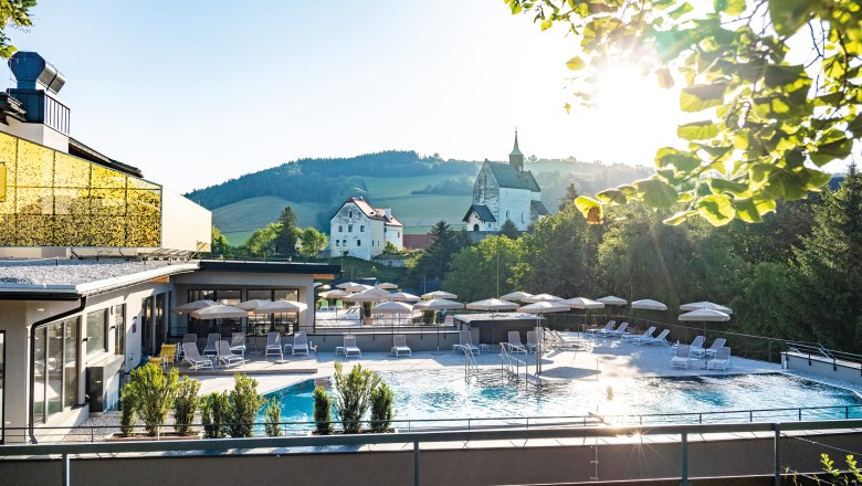Outdoor area of a hotel with pool, parasols and sun loungers, surrounded by nature and a church in the background.