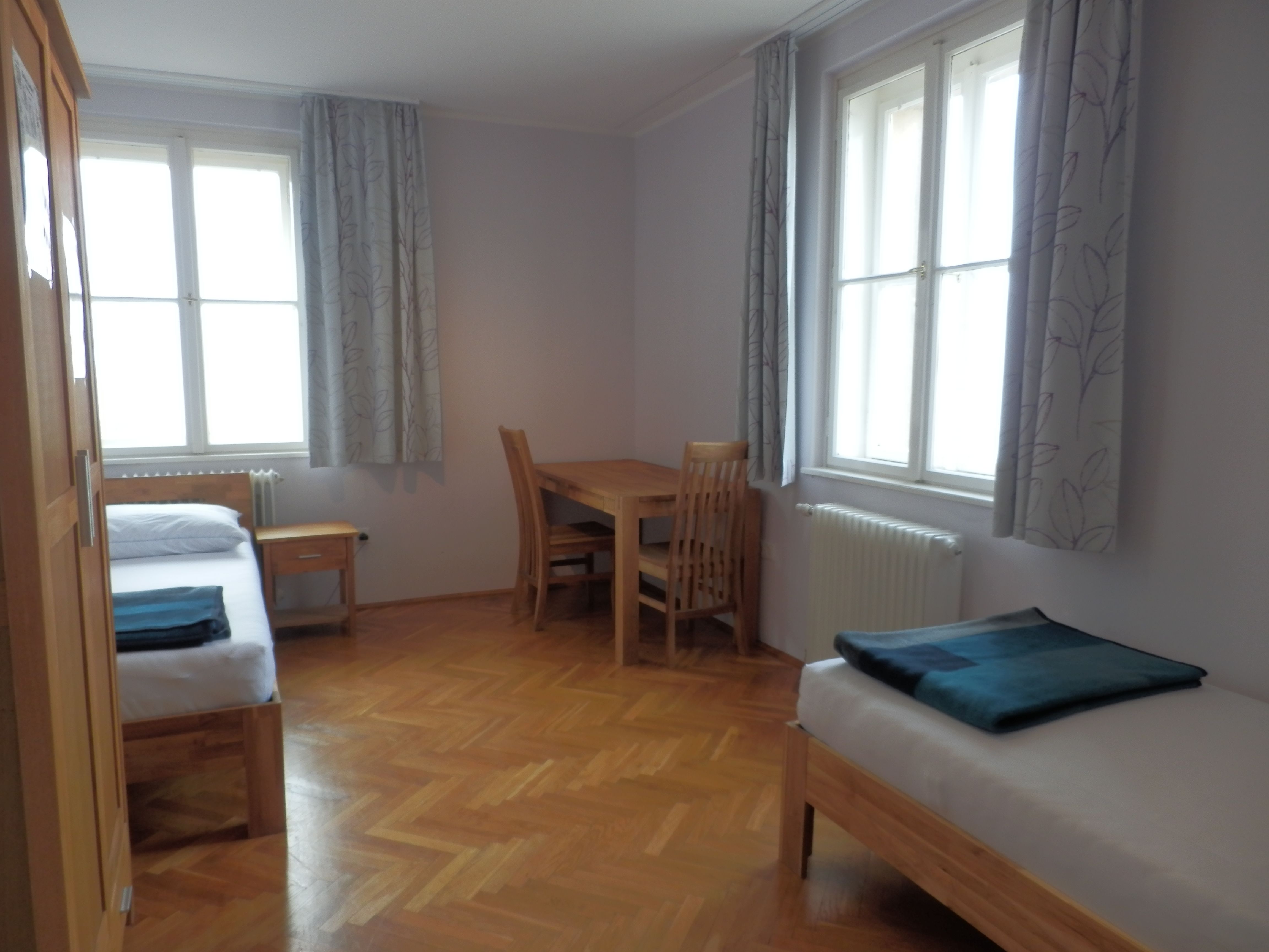 A simply furnished guest room with two beds, a table and chairs, wooden floor and large windows with curtains.