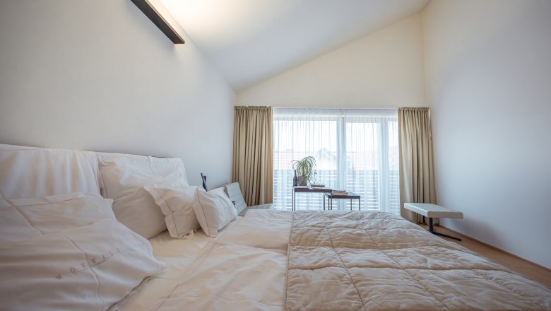 Modern hotel room with large bed, white pillows and bedspread, light-colored curtains and window in the background.