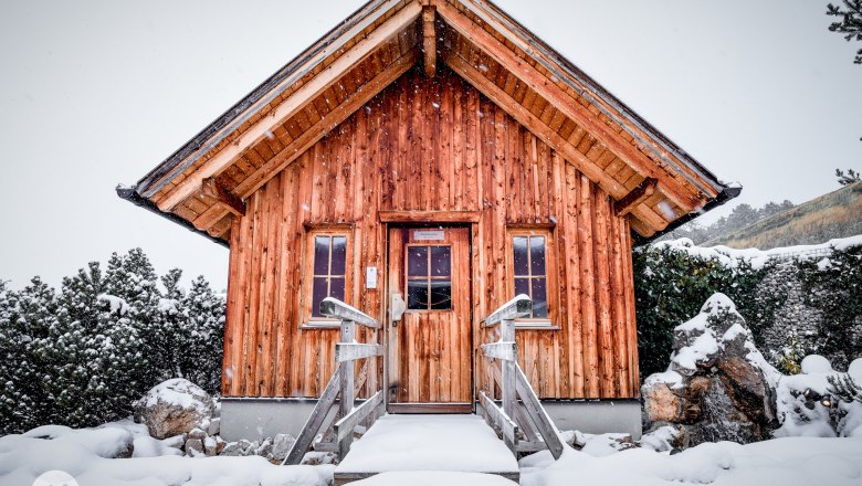 A small wooden hut in the snow with a steep roof and two windows.
