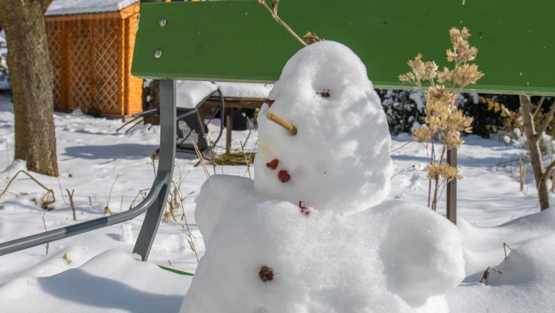 A little snowman sits on a green bench in the snow-covered garden.