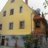 Yellow guest house with wooden veranda and flowering plants.