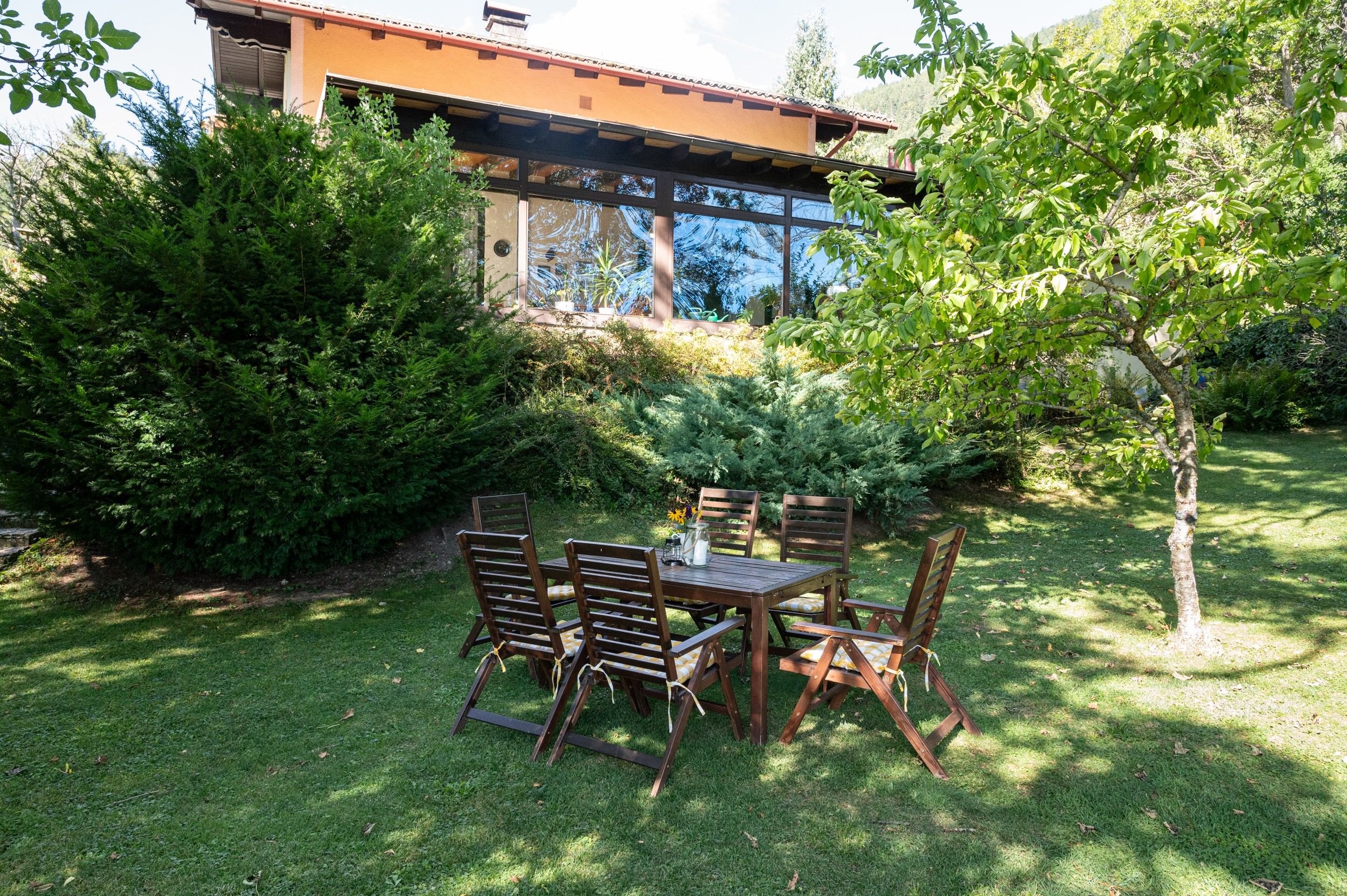 A garden with a wooden table and chairs in front of a vacation home with large windows and surrounded by trees and shrubs.