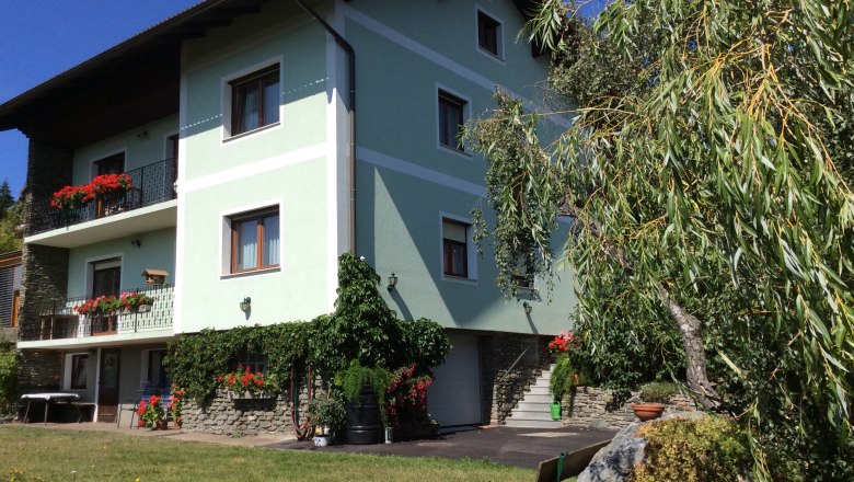 A green house with balconies full of flowers, surrounded by trees and a well-tended garden.