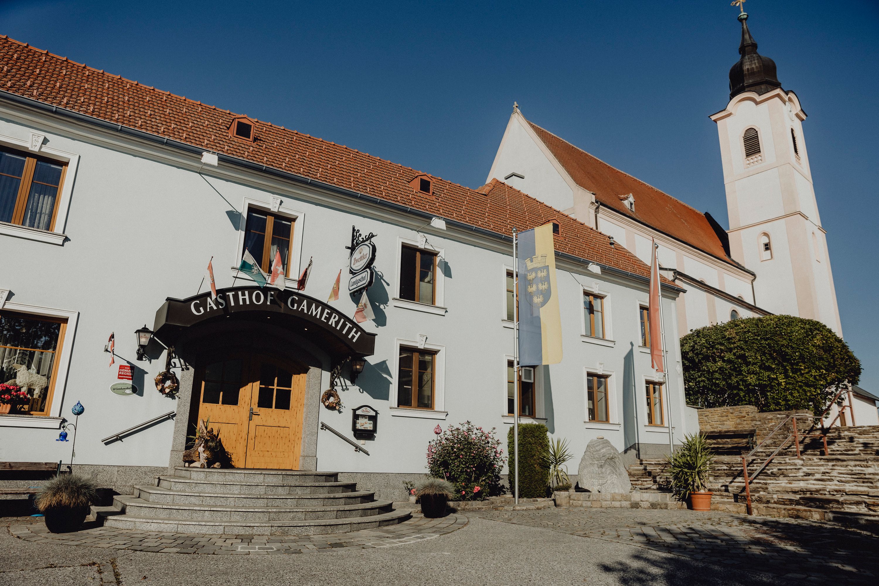 An inn with the inscription 'Gasthof Gamerith' next to a church with a tower under a blue sky.