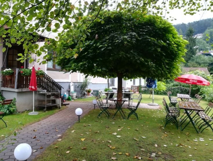 A cozy guest garden with tables and chairs under a large tree, surrounded by green countryside.