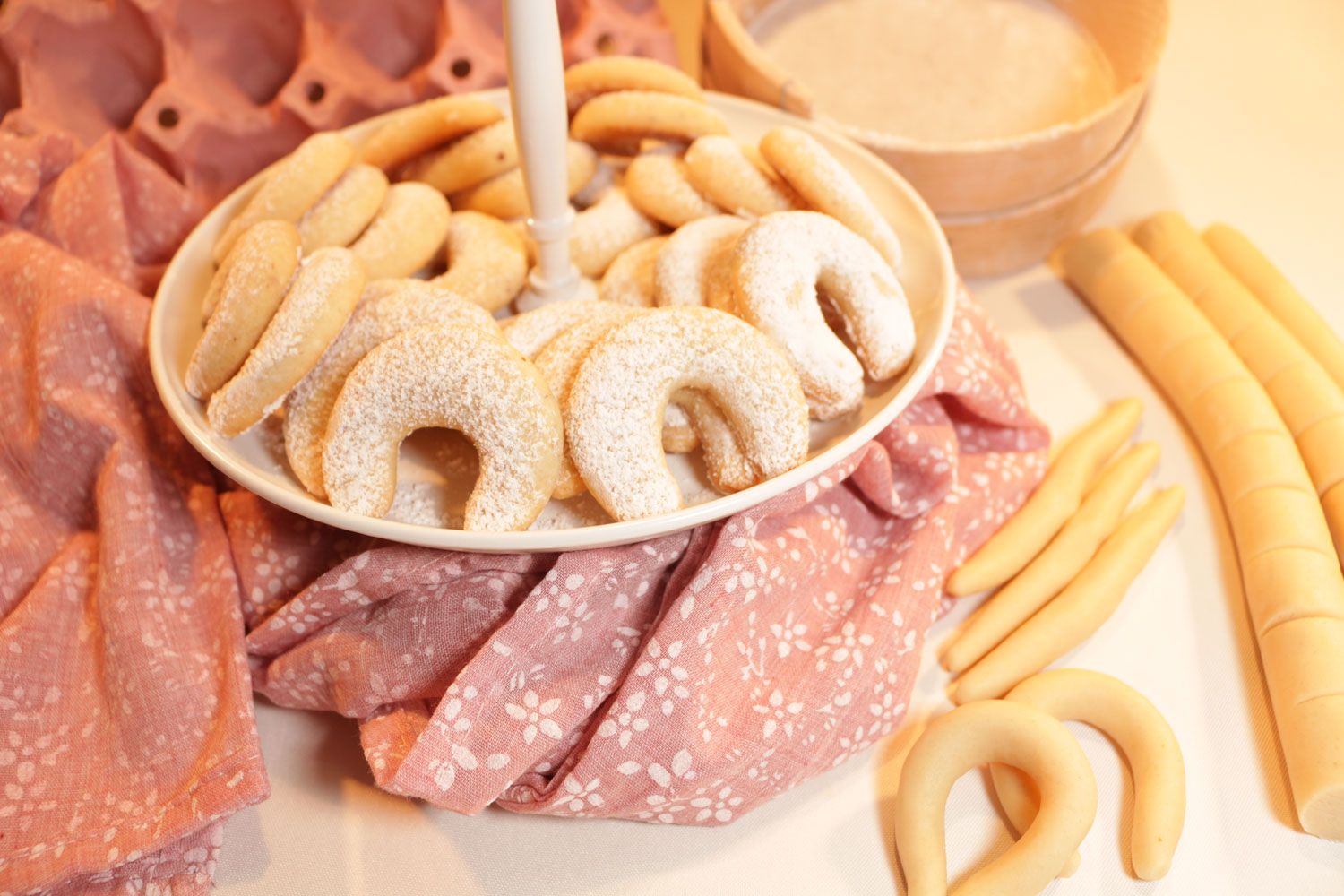 A plate of vanilla crescents on a pink cloth, surrounded by rolls of dough.