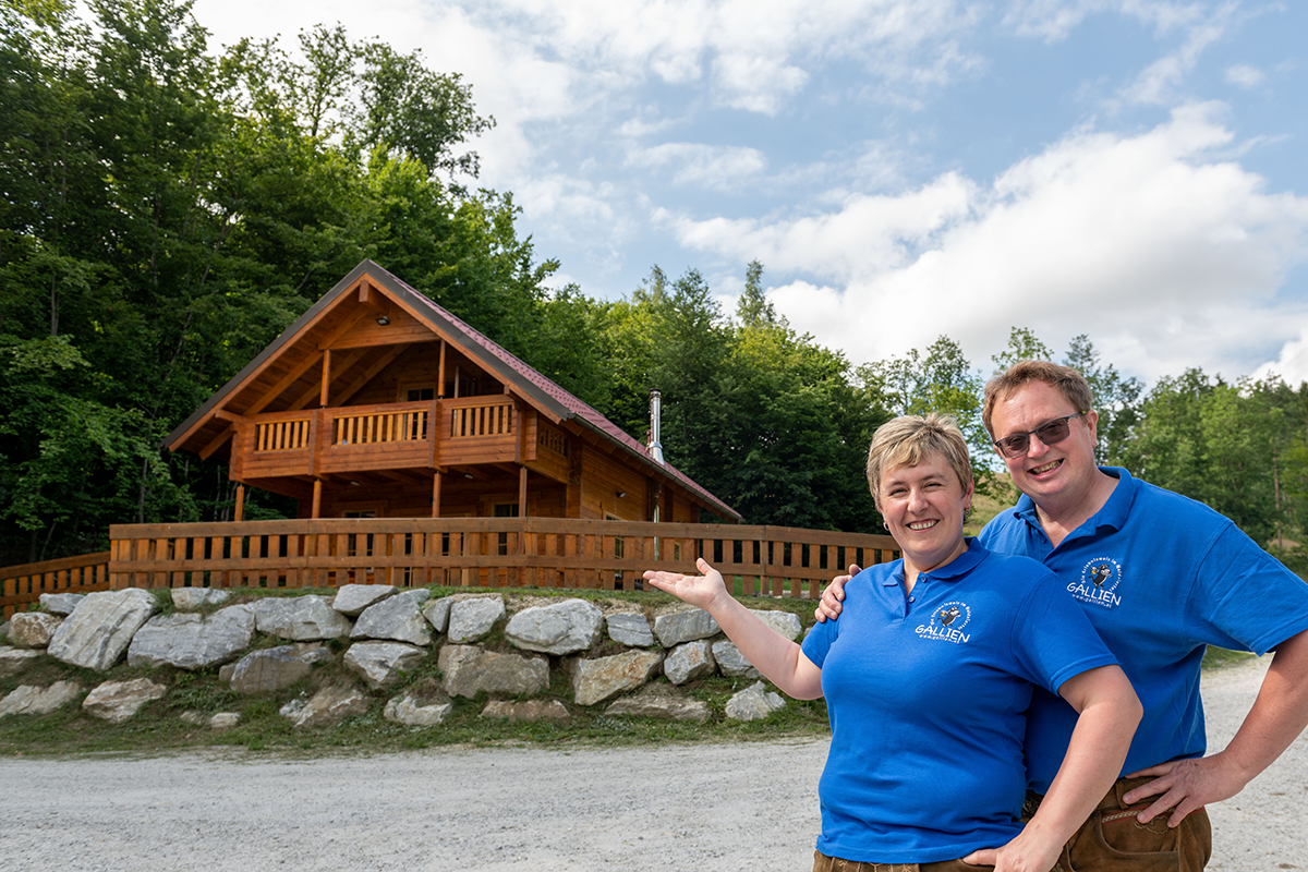 A couple wearing blue polo shirts is standing in front of a wooden cabin in a wooded area.