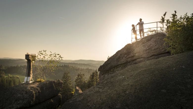 Two people standing on a rock with railings, sunset and forest in the background.