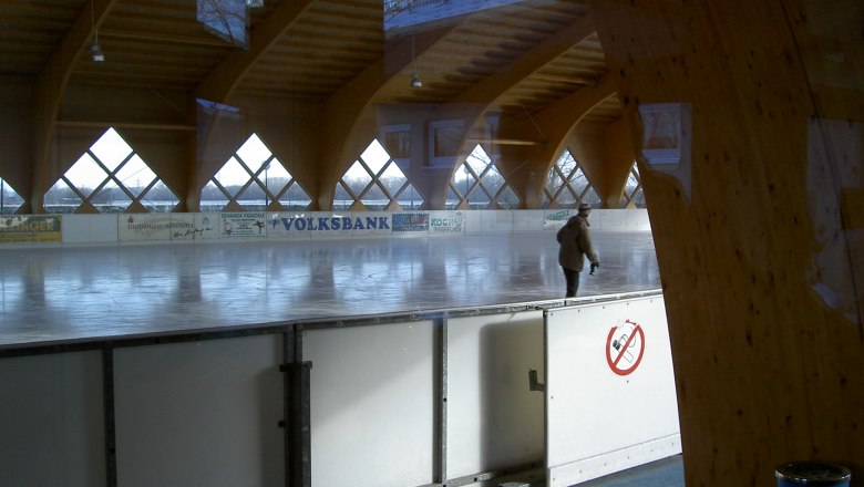 A covered artificial ice rink with a person skating on it. The rink is surrounded by wooden beams and windows.